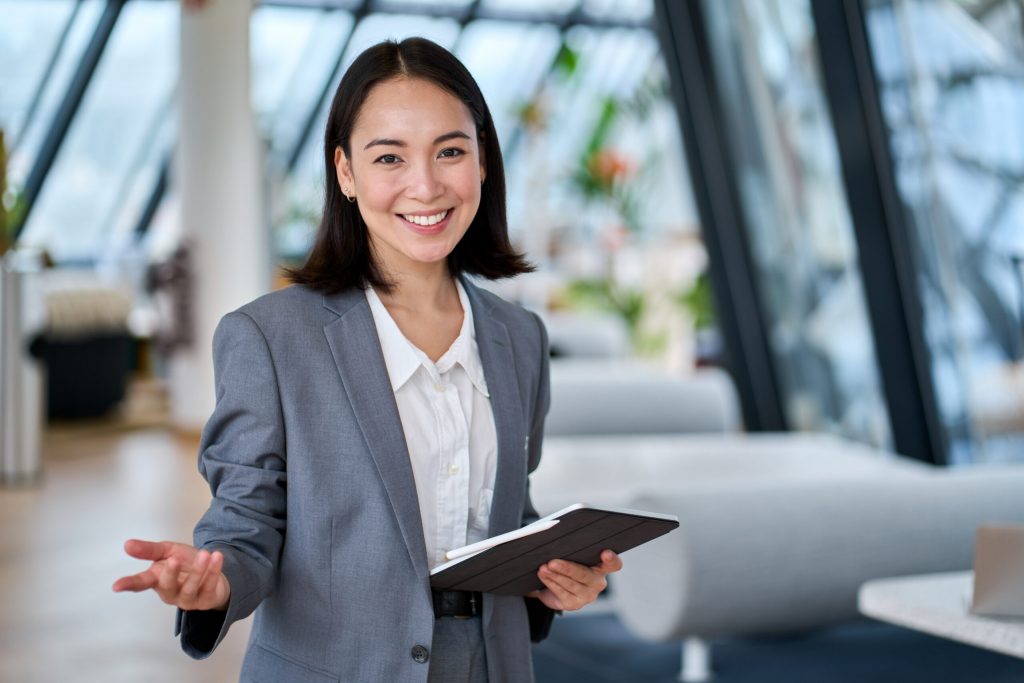 Smiling woman holding a clipboard, welcoming the viewer in a modern office setting